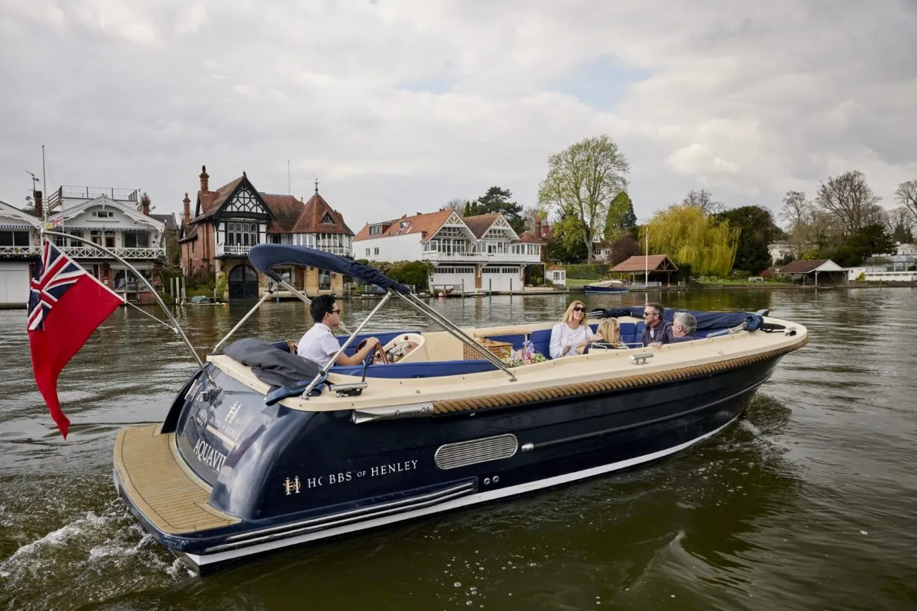 Hobbs of Henley boat Aquavir cruising along the river thames, driven by a skipper with 4 passengers on board. The sky is cloudy, and there are houses pictured in the background that are right on the river.